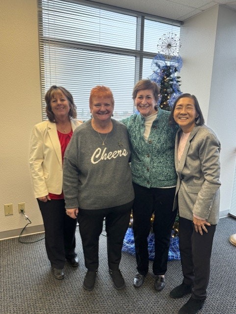 Four women are smiling together in an office setting with a decorated Christmas tree in the background.