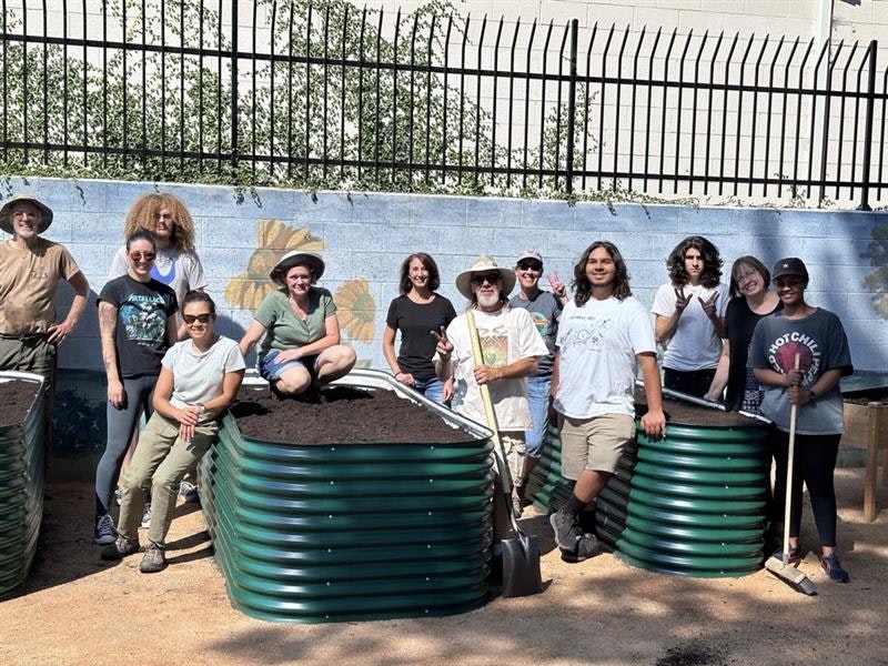 A group of people poses with gardening tools around raised garden beds filled with soil, against a mural-covered wall.