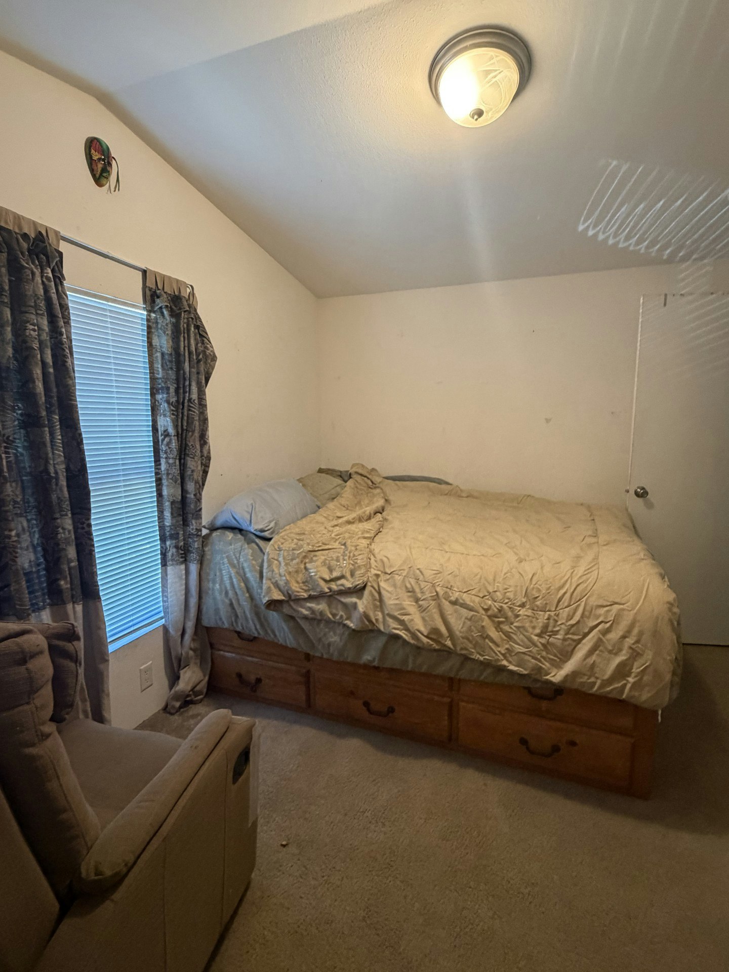 A cozy, simple bedroom with a bed, a chair, curtains, and a light fixture; features a decorative mask on the wall.