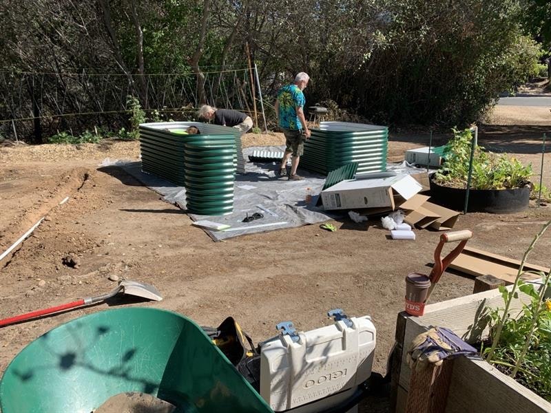 People assembling green corrugated garden beds outdoors, surrounded by tools, boxes, and plants.