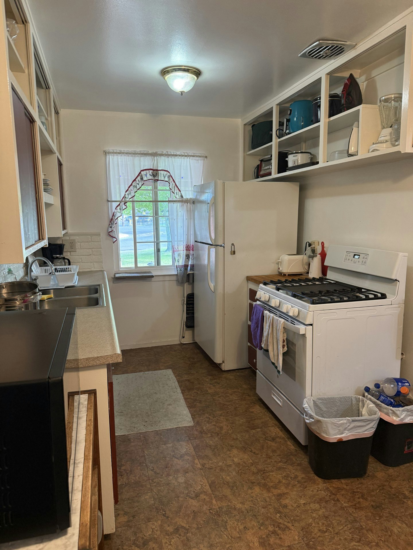 A small, tidy kitchen featuring a stove, fridge, microwave, and shelves with various appliances and utensils.