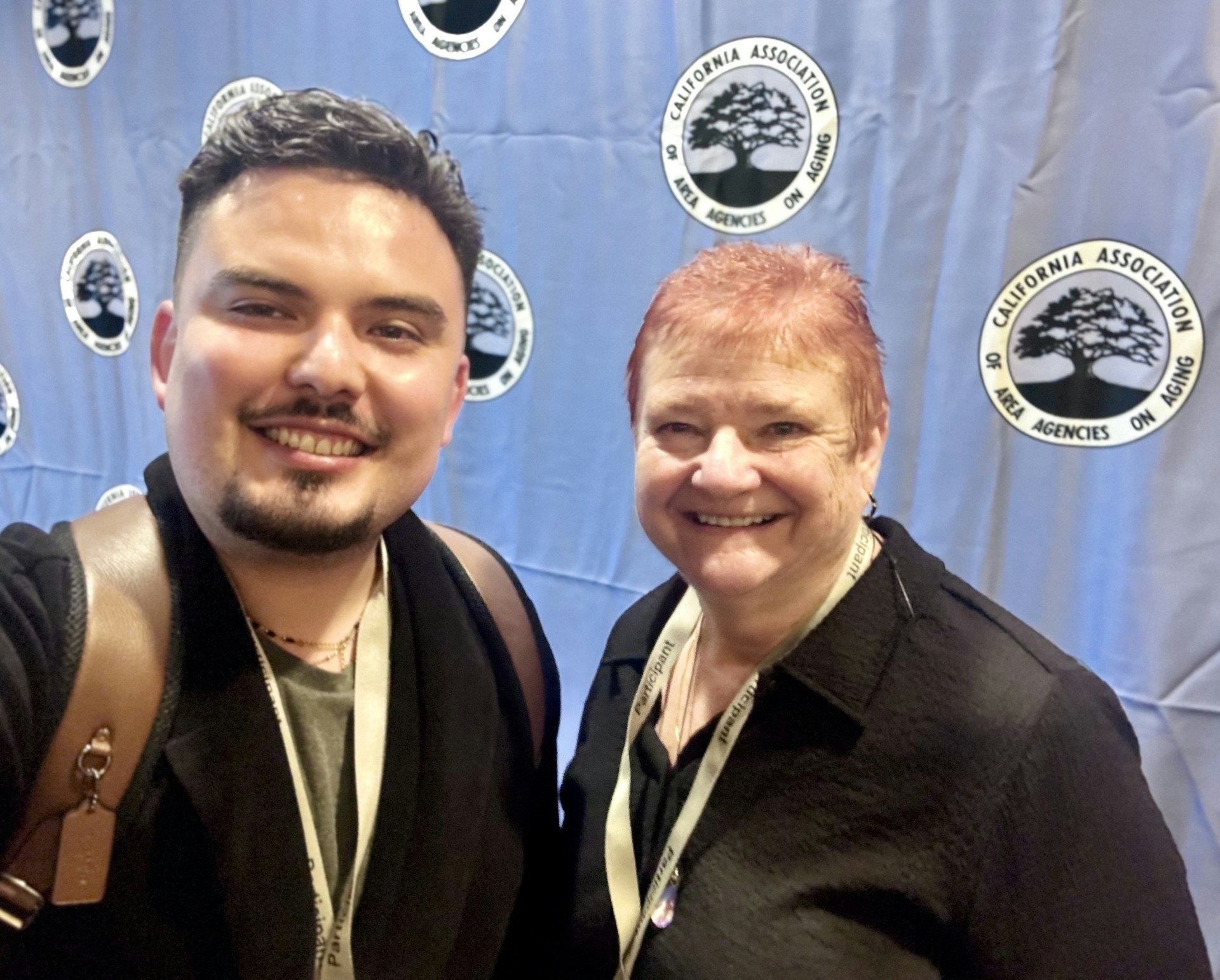 A smiling man and woman pose for a selfie in front of a backdrop featuring the California Association of Area Agencies on Aging.