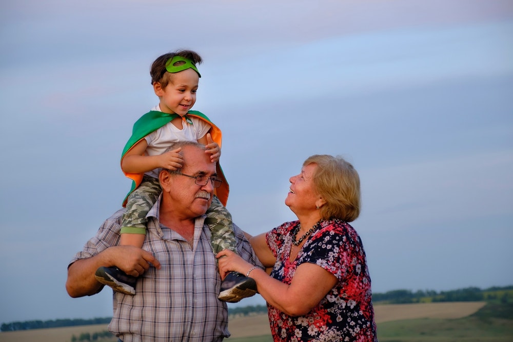 A joyful child dressed as a superhero sits on a man's shoulders while a woman smiles at them outdoors.
