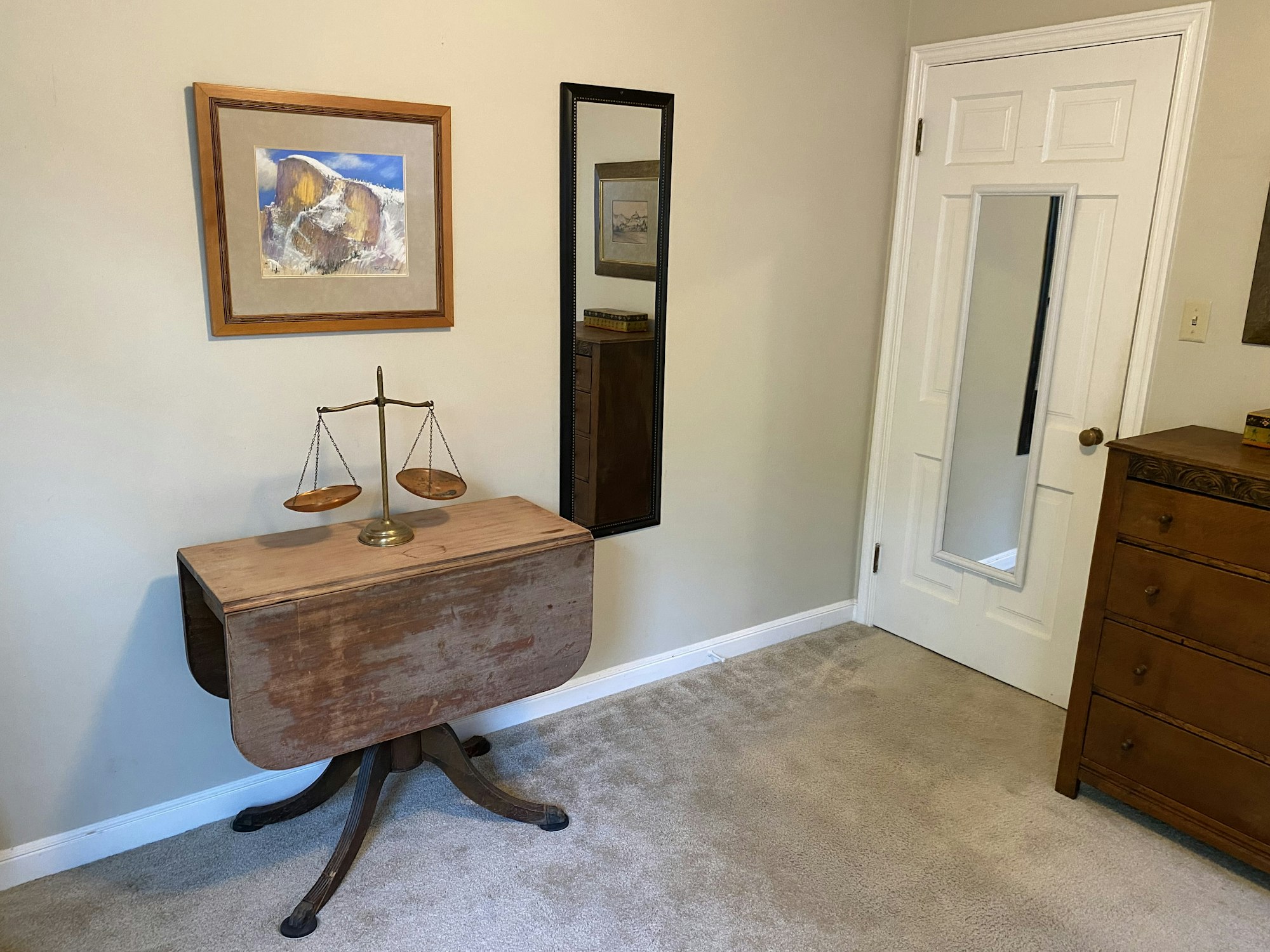 A small room with a wooden table, scales, framed artwork, mirror on a door, and a dresser on carpeted floor.