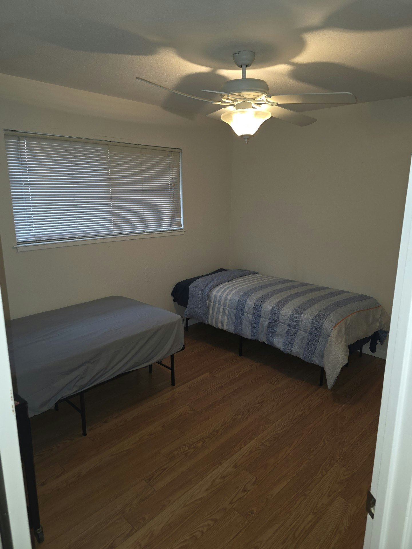 A simple bedroom with two beds, a ceiling fan, and a window with blinds. Clean, minimalistic design.