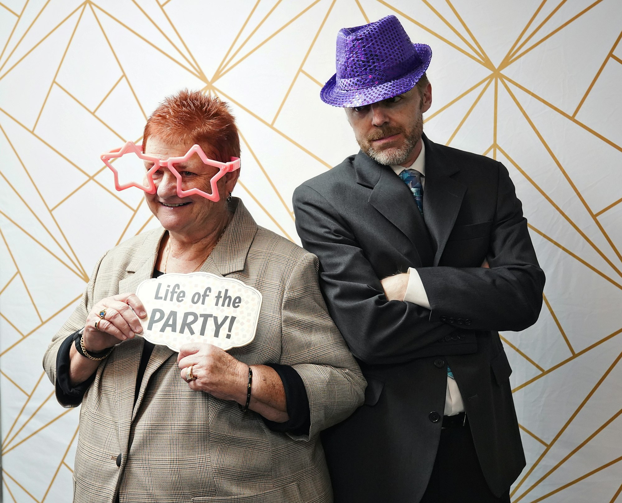 A woman with star-shaped glasses and a sign says "Life of the PARTY!" stands next to a serious man in a suit and purple hat.