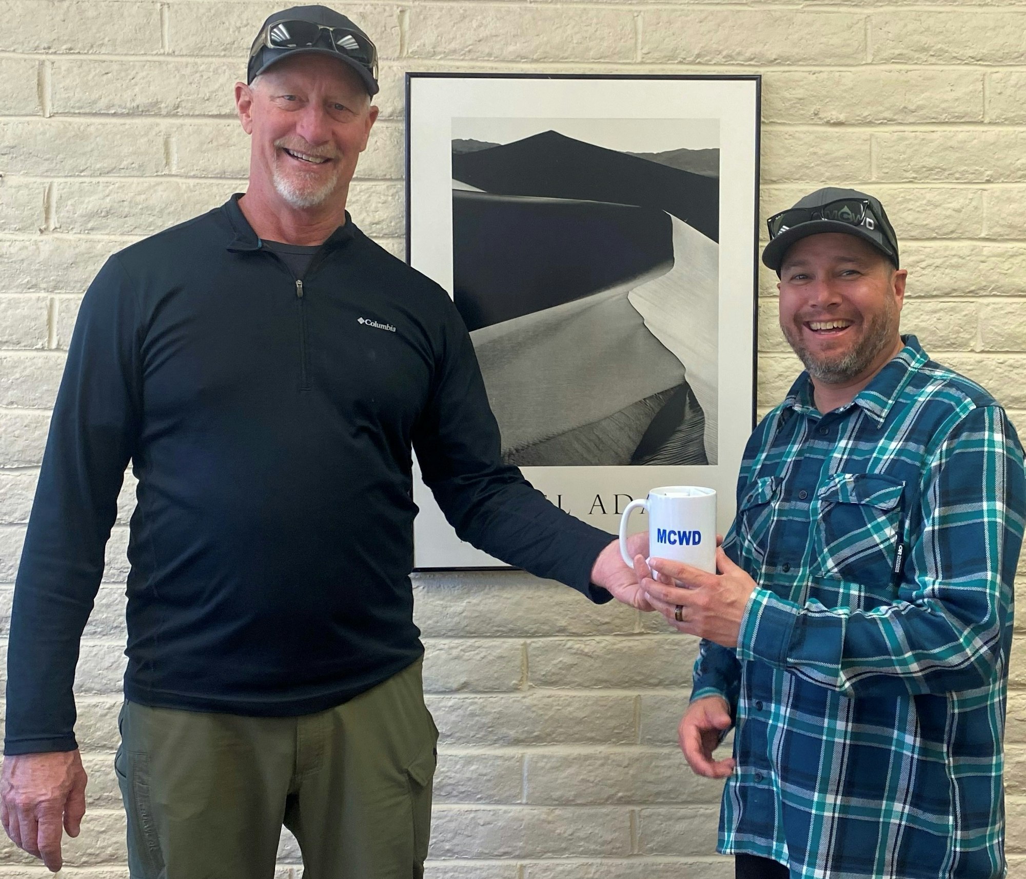 Two men are smiling and exchanging a mug that says "MCWD" in front of a photo of dunes on a brick wall.