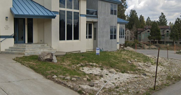 Modern house with a blue roof, large windows, and a "For Sale" sign on a rocky landscape.