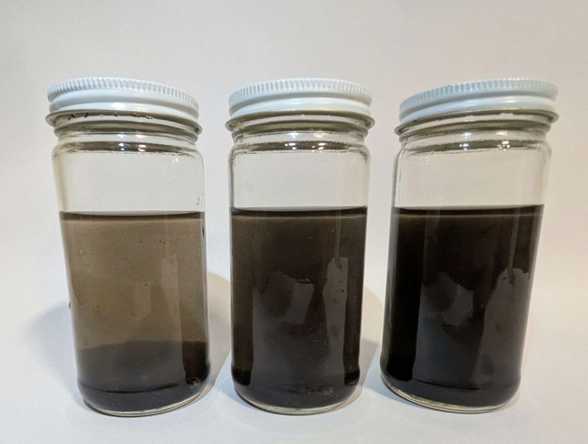 Three glass jars with white lids, each containing different shades of dark liquid.