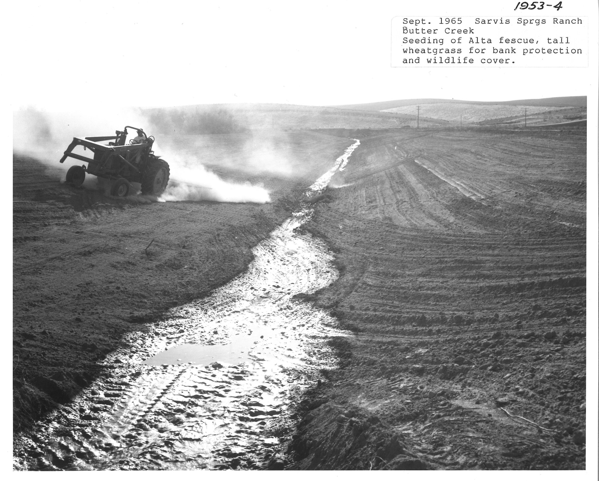 A tractor works on a dirt field beside a water channel; 1965 Sarvis Springs Ranch.