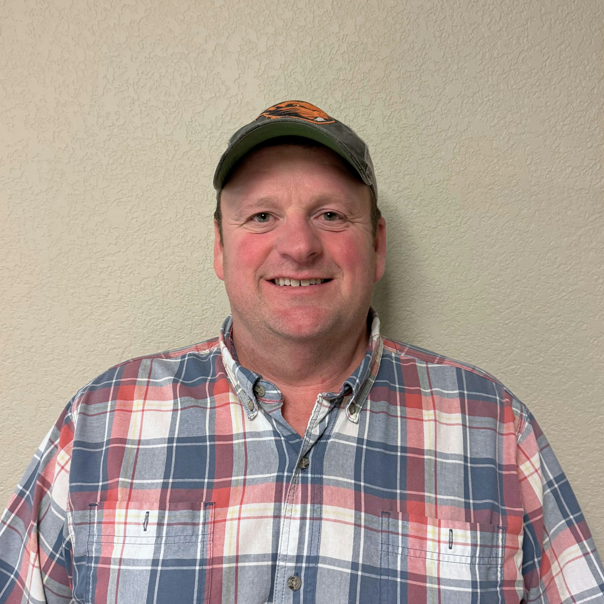 A man wearing a plaid shirt and a cap, standing against a textured wall, smiling at the camera.