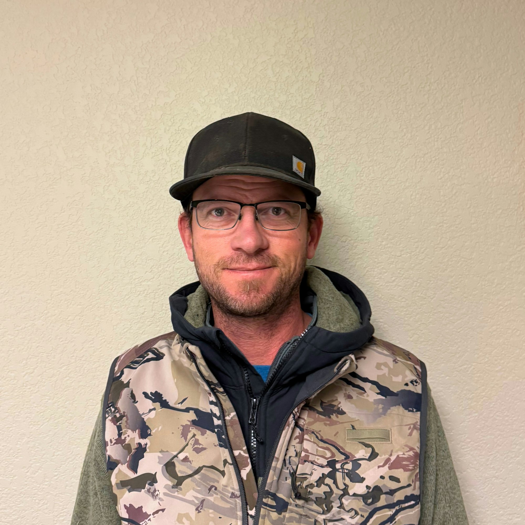 A person wearing a camo vest, hoodie, cap, and glasses, standing against a plain wall.
