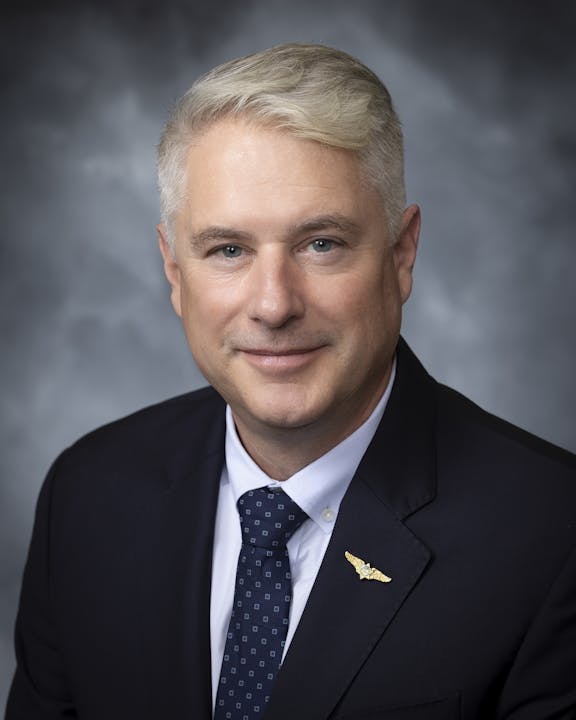 Portrait of board member John Gaglioti smiling in a blue suit and tie against a gray background