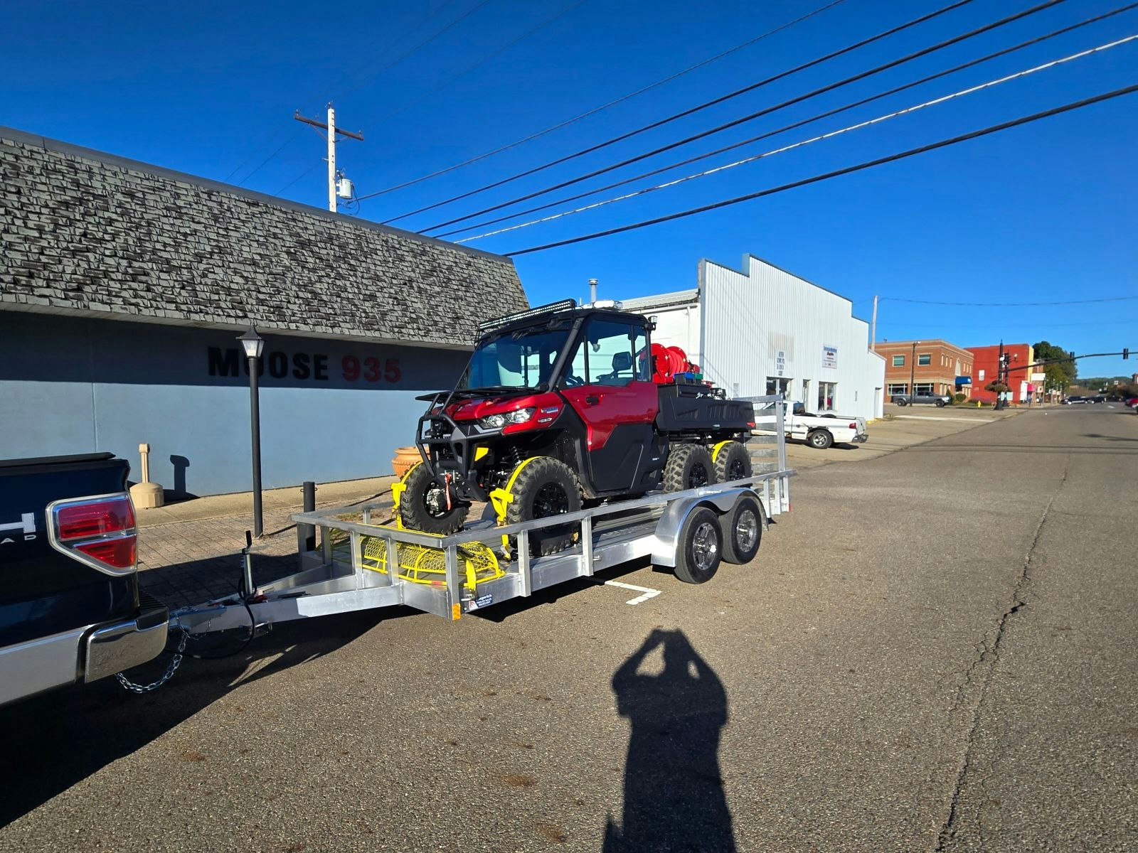 A red utility vehicle on a trailer hitched to a pickup truck, with urban buildings in the background and power lines overhead.