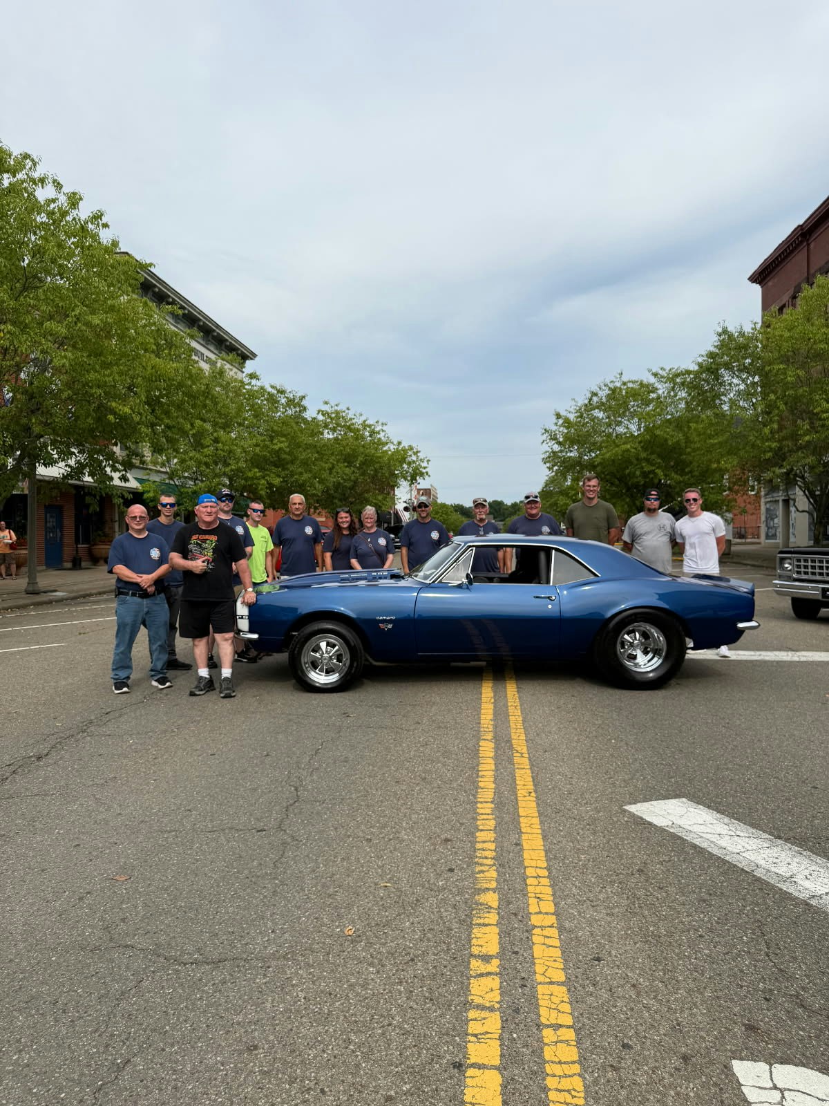 A group of people standing behind a blue vintage car on a street with trees and buildings in the background.