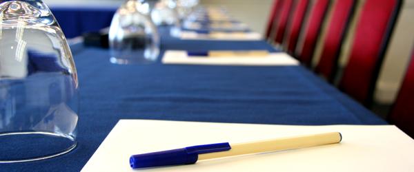 A conference table setup with pens, papers, and inverted water glasses against a background of red chairs.