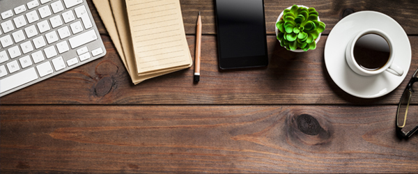 A keyboard, notebook, pen, smartphone, plant, coffee cup, and glasses on a wooden desk.