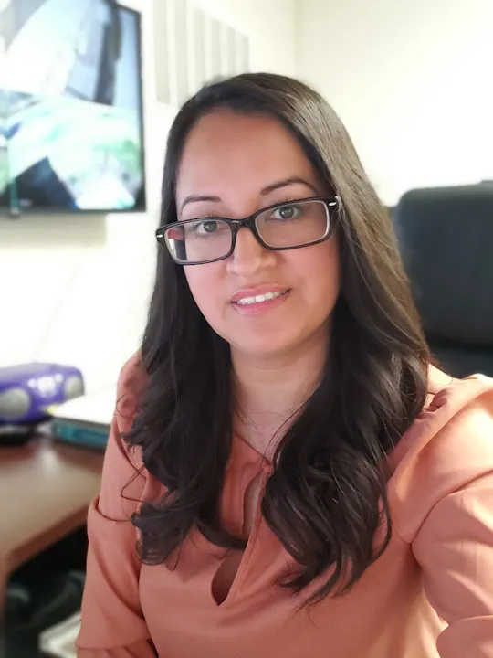A person with long hair and glasses, wearing a rust-colored top, is sitting indoors.