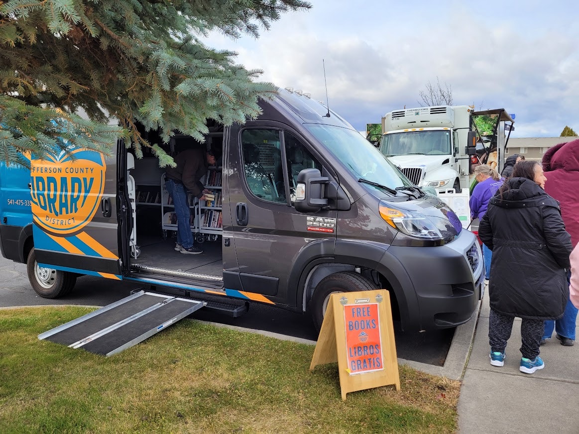 A library truck offers free books, with people browsing the selection and a nearby sign promoting the giveaway.