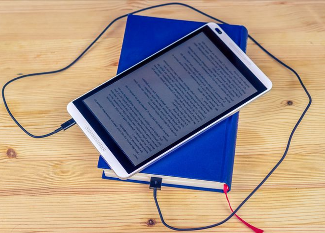 An eBook reader sitting on top of a print book and connected by a charging cord