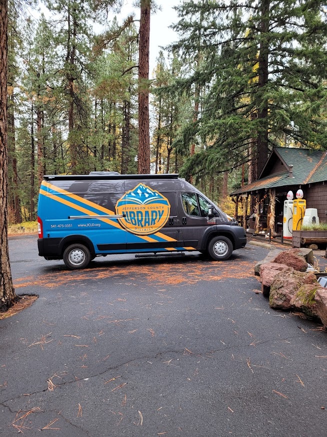 A library van from Jefferson County is parked in a wooded area, alongside a small cabin and some rocks.