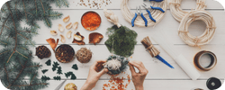 Crafting materials on a table with hands making a decoration; includes greenery, ribbons, and seasonal items.