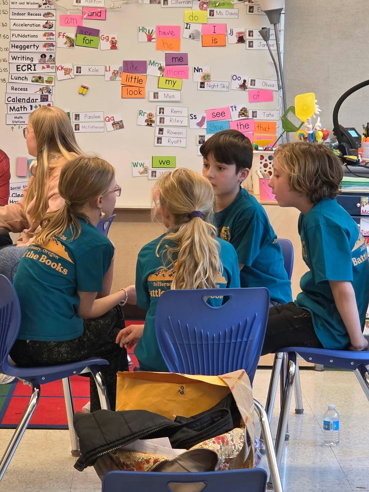 A group of children in teal shirts discuss together in a classroom with colorful posters on the wall behind them.