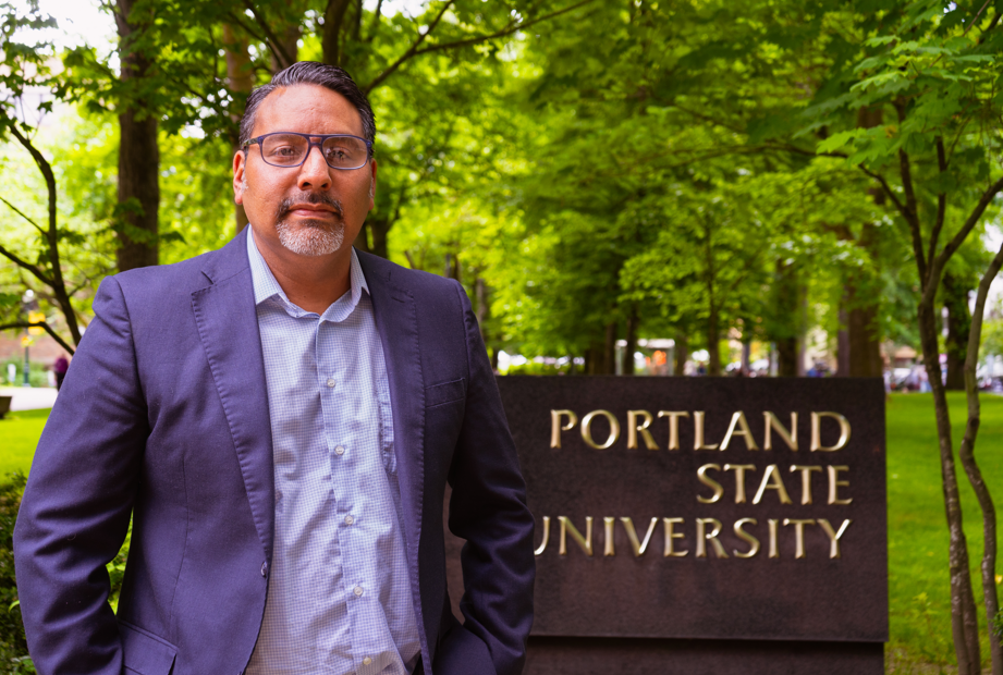 A man in business attire stands in front of a sign for Portland State University, surrounded by greenery.