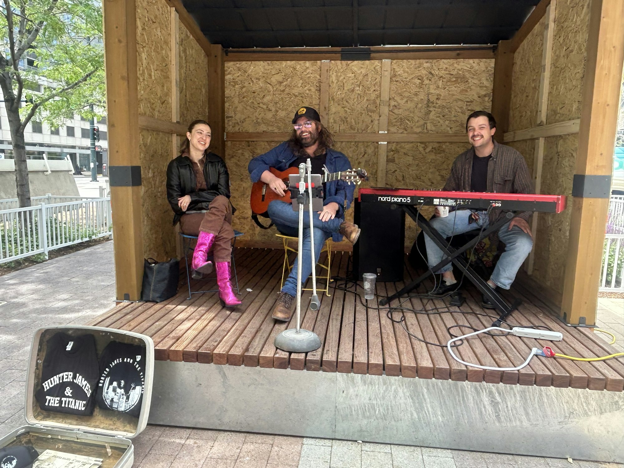 Three people in a wooden booth: one with a guitar, another at a keyboard, and one sitting. An open case displays band merch.