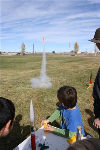 A small rocket is launching in a grassy field as children watch.
