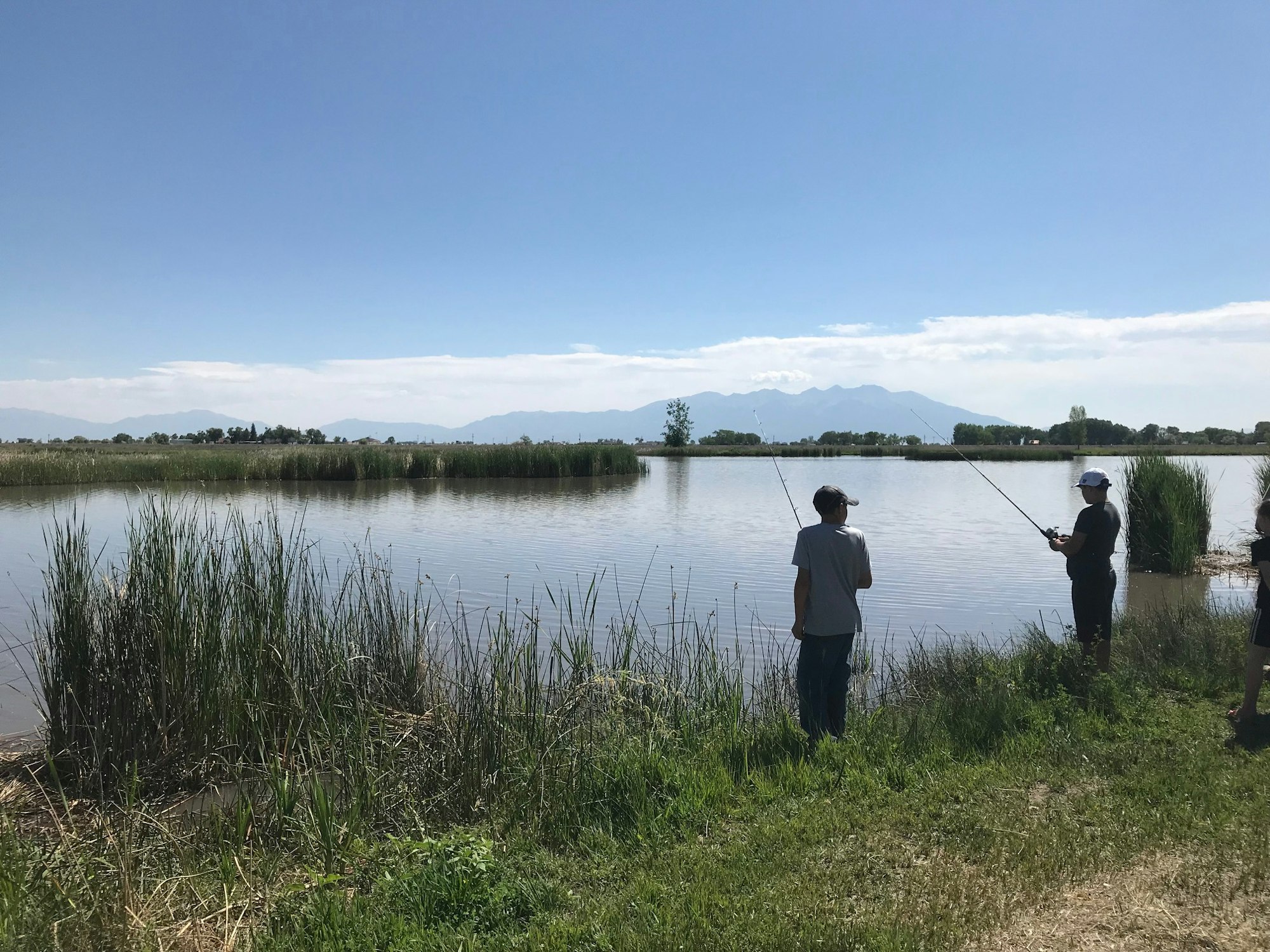 Two people fishing by a lake with mountains in the background and a clear blue sky.