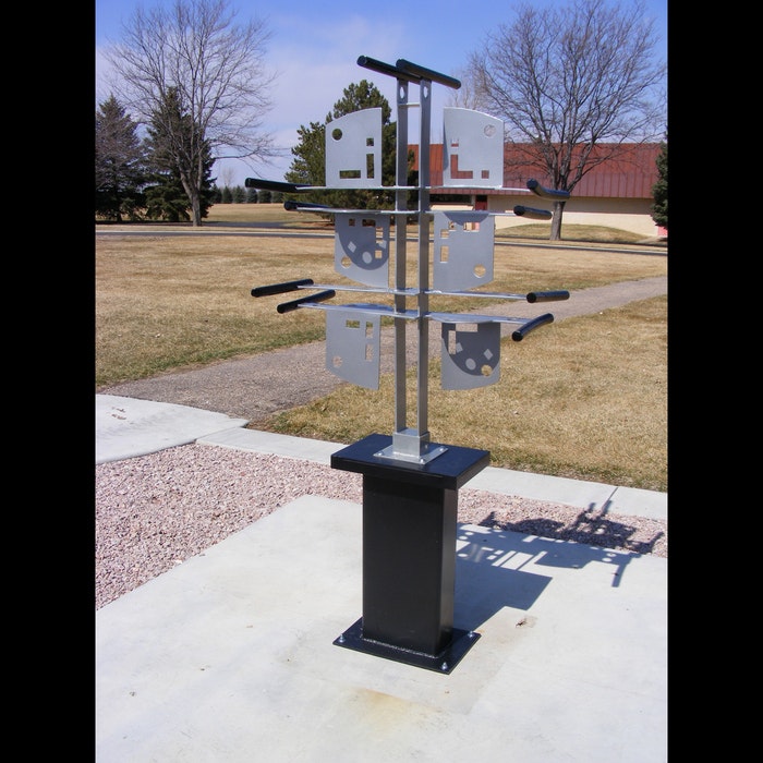 A metallic wind sculpture with rotating blades mounted on a pole, situated in an outdoor park setting.