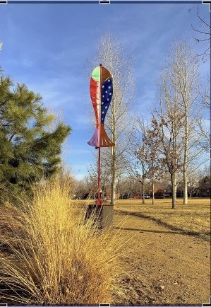 A colorful rocket-shaped sculpture with star patterns stands in a park, surrounded by trees and dry grass under a blue sky.