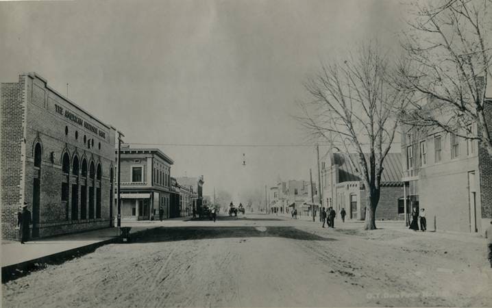 A vintage street scene with people walking, horse-drawn carriages, and historic brick buildings on both sides. Trees line the street.