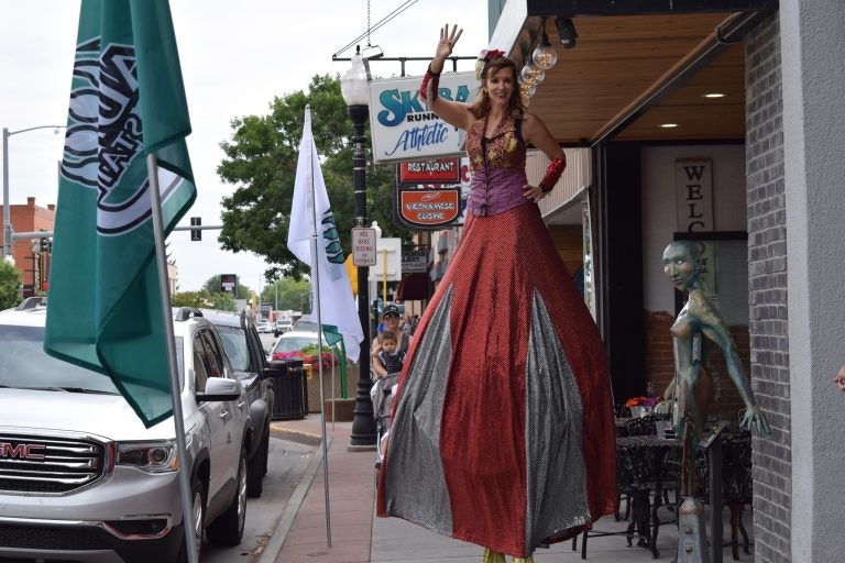A person on stilts in a long red dress waves on a sidewalk next to an alien statue, with cars and signs in the background.