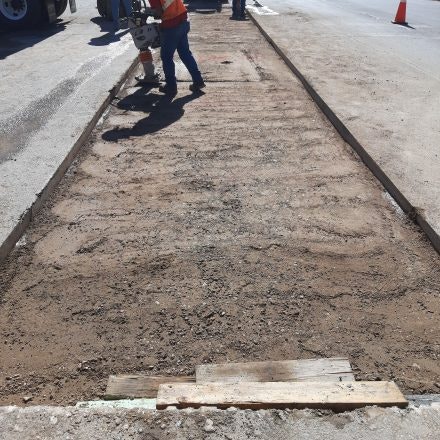 A person in a safety vest is using equipment on a patched-up road section, with traffic cones nearby.