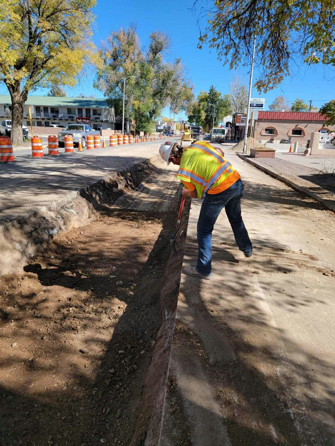 Construction worker digging a trench on the roadside with orange barriers in the background.