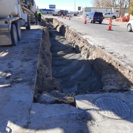 Road construction with a trench, workers, and traffic cones; cars and a mixer truck nearby.