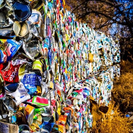 Stacks of compressed aluminum cans and packaging materials in an outdoor recycling area with trees in the background.