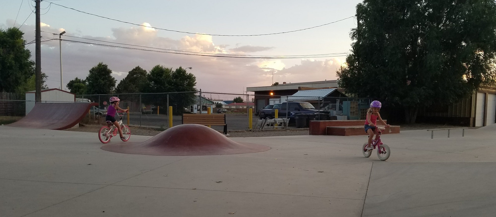 Two children wearing helmets are biking at a skatepark during sunset.