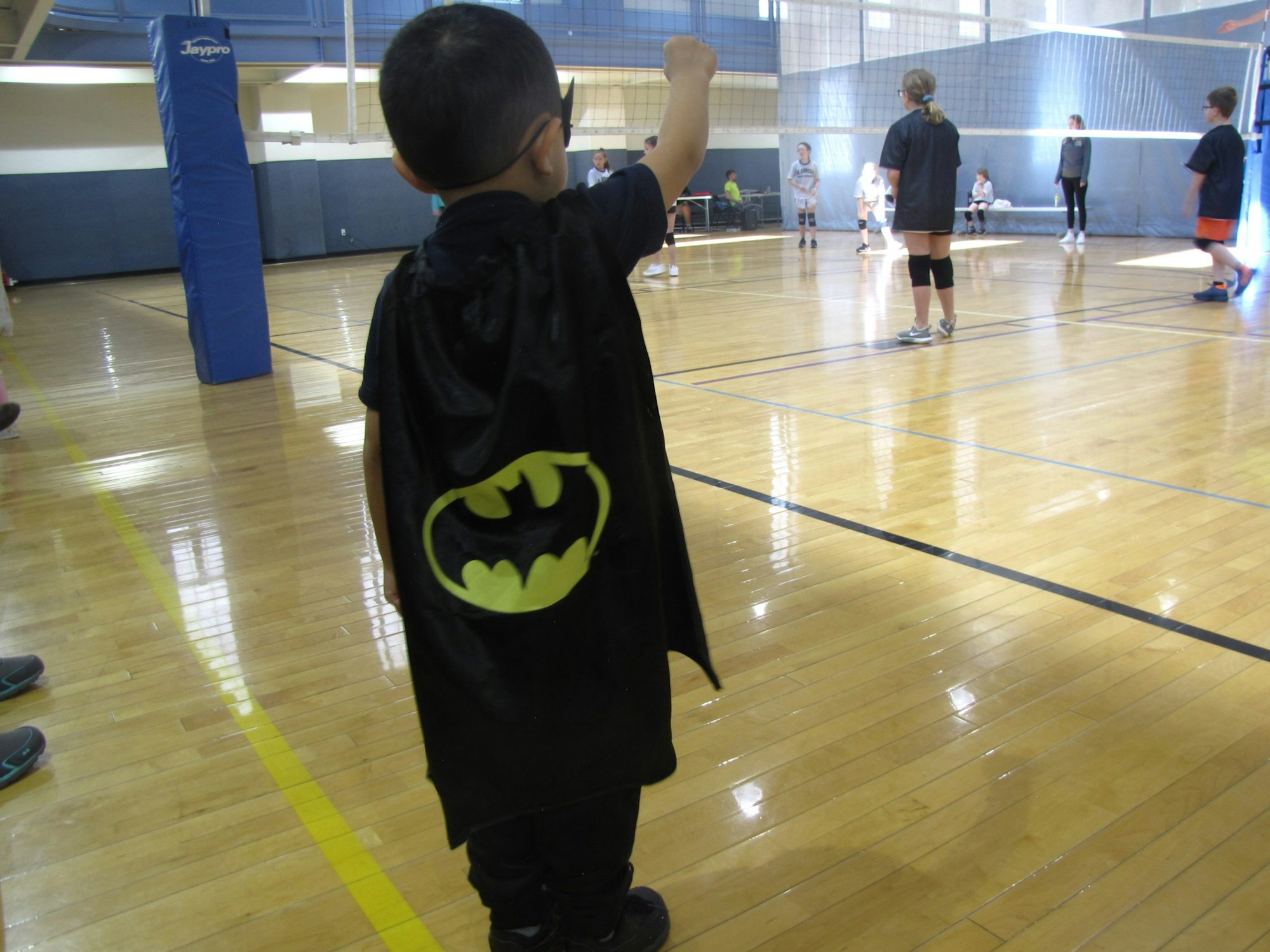A child wearing a Batman cape stands on a gym floor near a volleyball game, raising one arm.