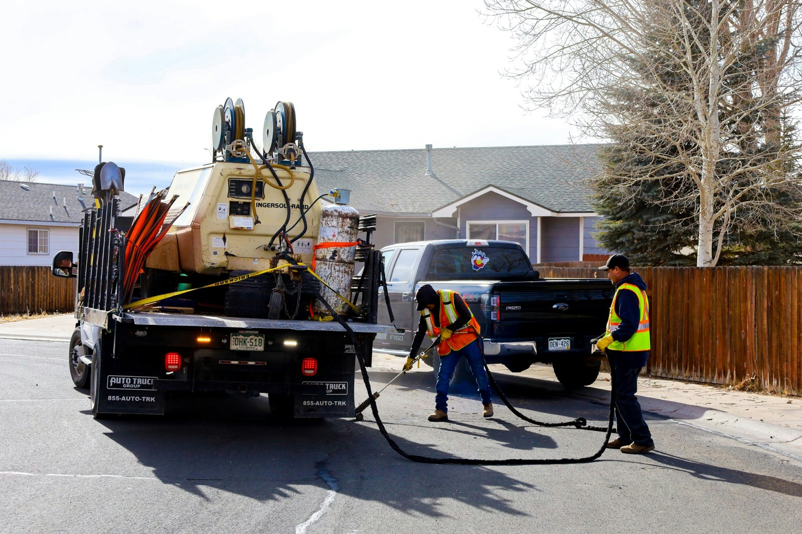 Two workers in safety vests fill asphalt cracks using equipment from a truck in a residential area.