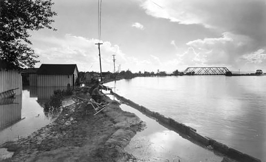 Flooded area with partially submerged houses, sandbags along a path, and a metal bridge in the distance under a cloudy sky.