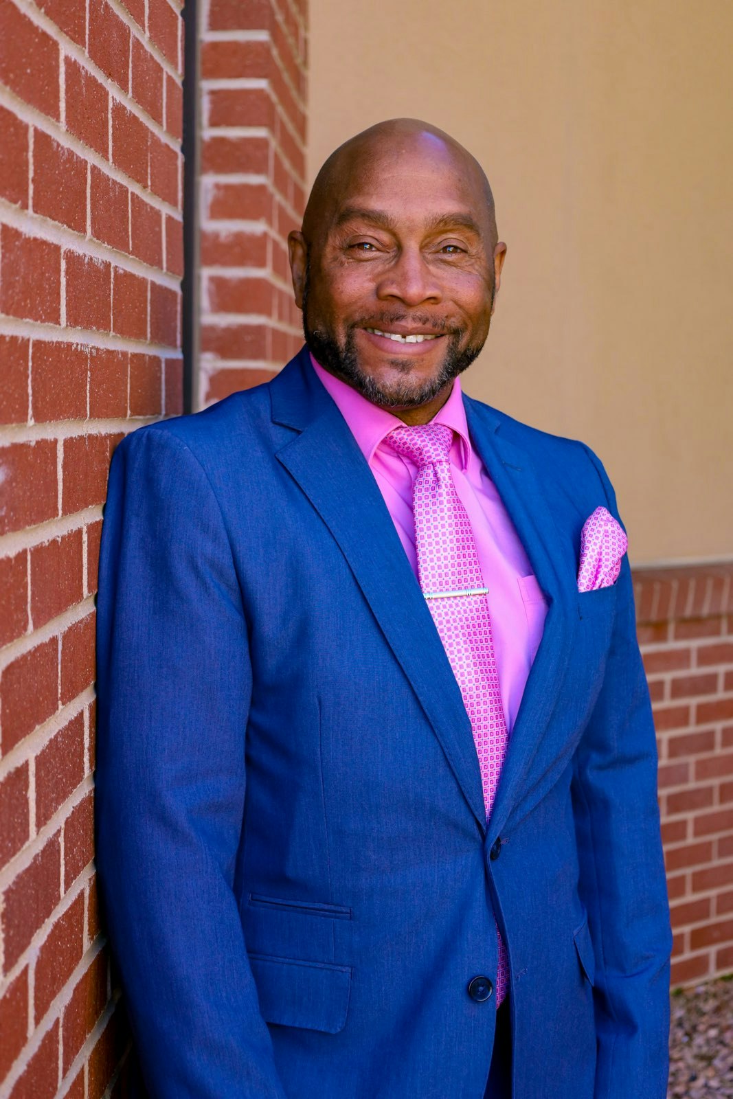 Smiling man in a blue suit with a pink shirt and tie, standing against a brick wall.