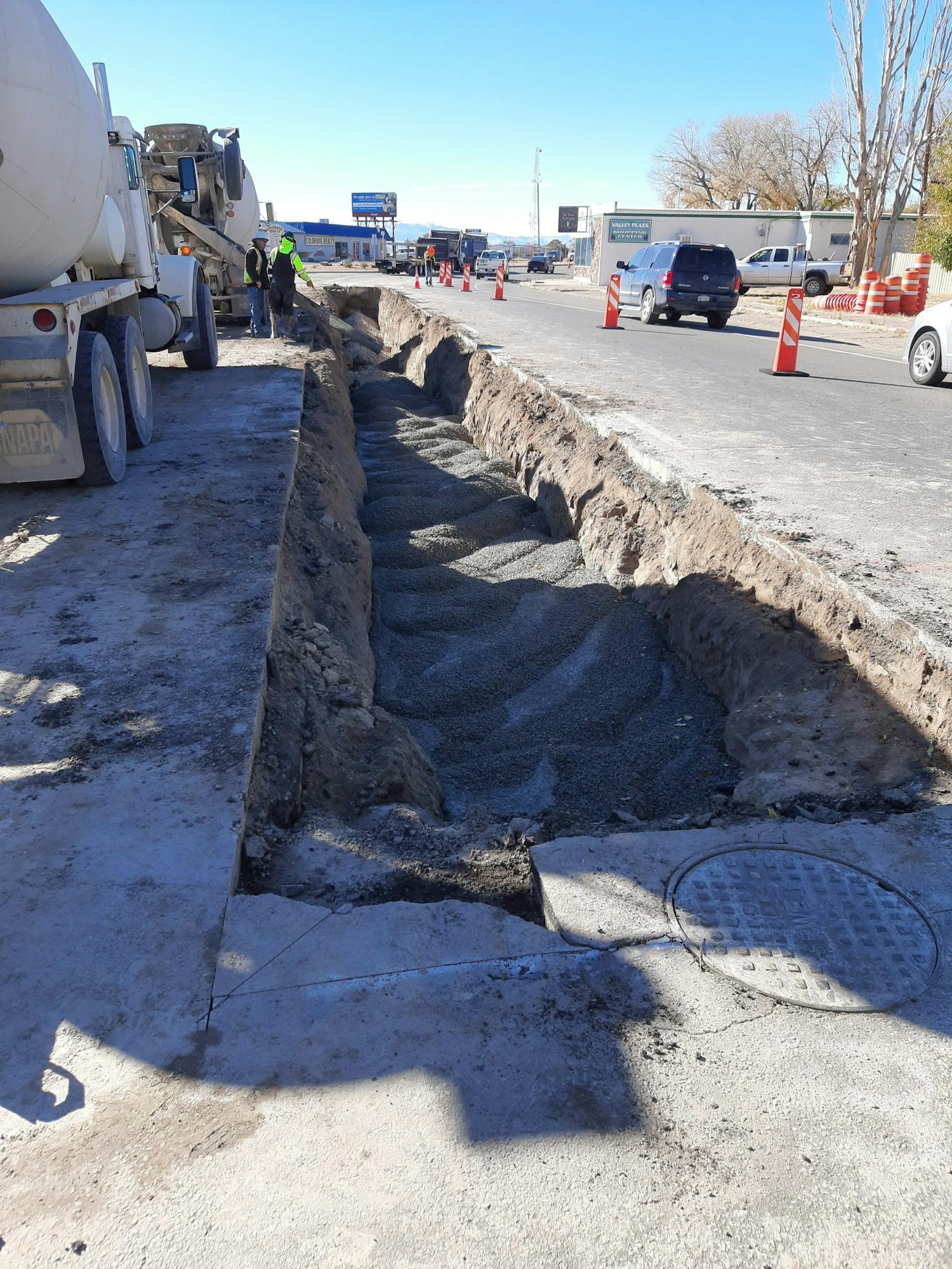 Construction site with a trench being filled with concrete, workers, traffic cones, and vehicles on the road.