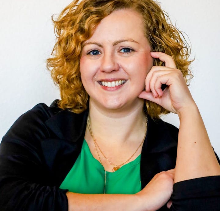 A person smiling, with curly hair, wearing a green top and a necklace, against a plain background.