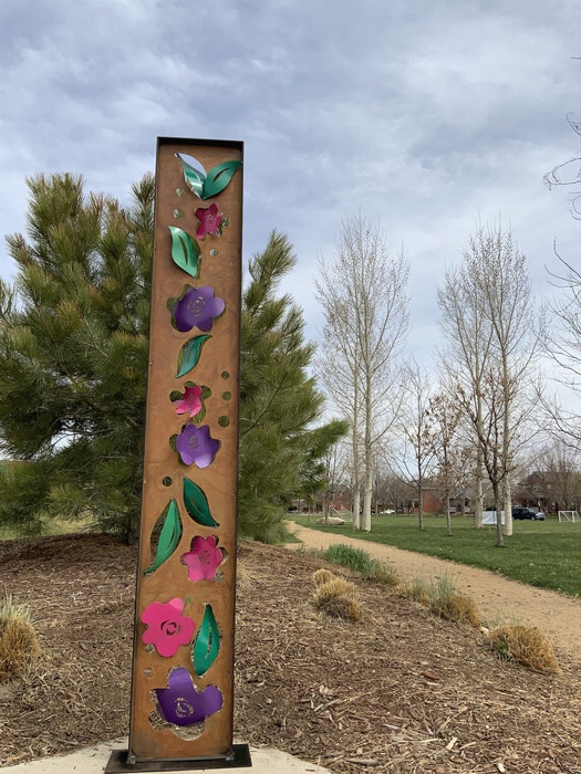 A tall metal sculpture with colorful flowers and leaves, set outdoors near trees and a walking path.