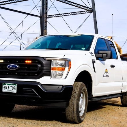 A white Ford truck with a Colorado license plate, labeled "Alamosa," parked on gravel near a metal structure.