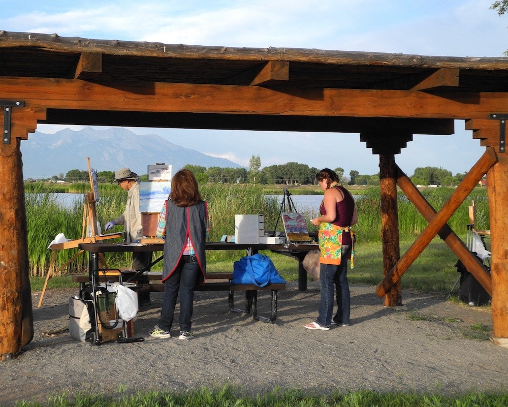 People painting outdoors under a wooden shelter, with mountains and a lake in the background.
