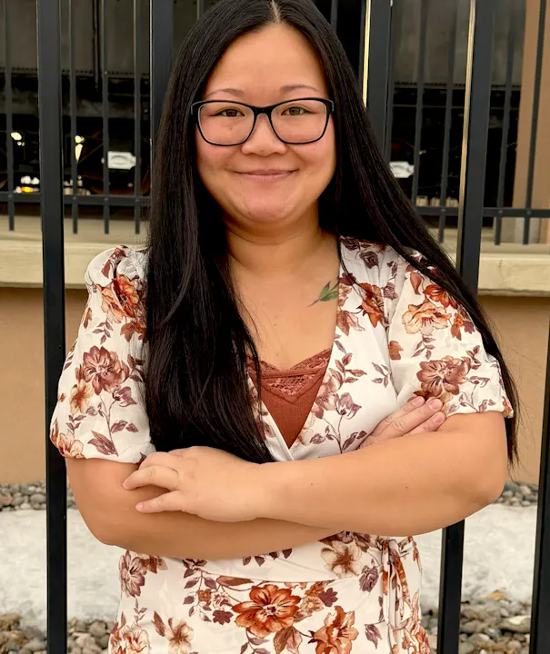 A woman with long dark hair and glasses stands confidently with arms crossed, wearing a floral-patterned dress against a backdrop of a fence.